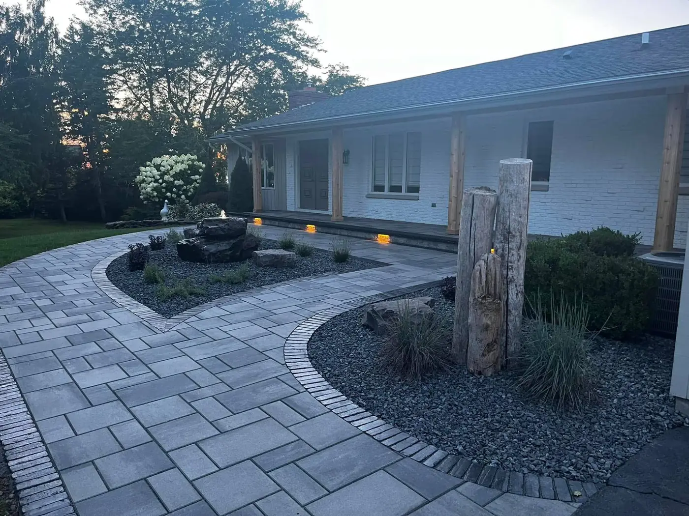 Paver walkway and front porch at dusk with a stone fountain, landscape beds and step lights