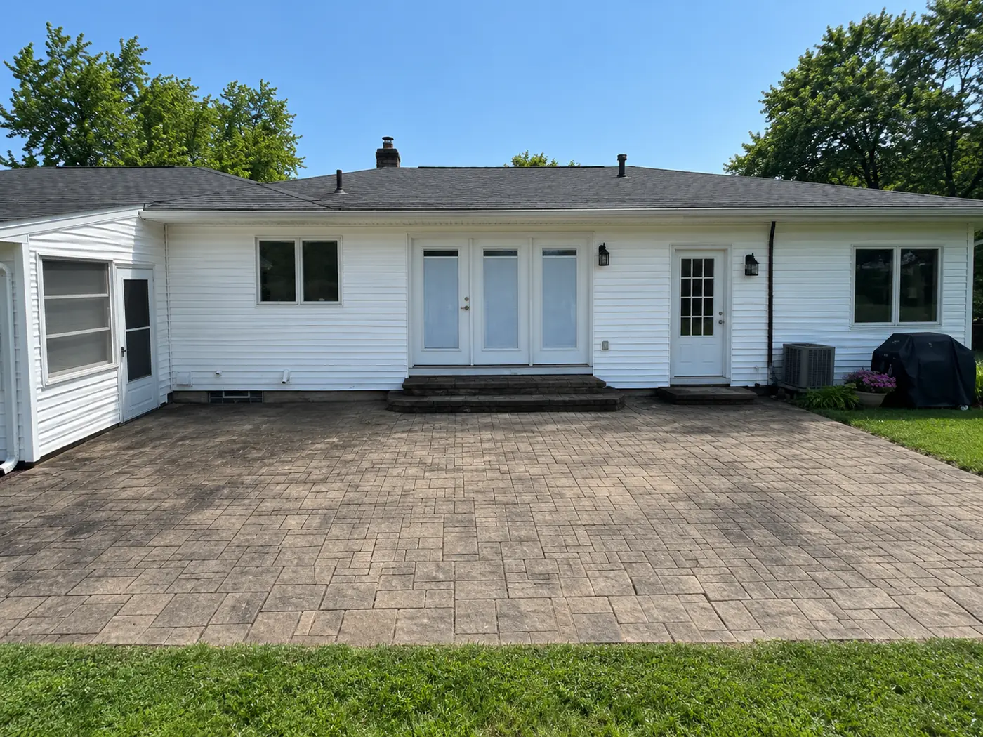 Dirty, stained paver patio behind a white ranch home before maintenance