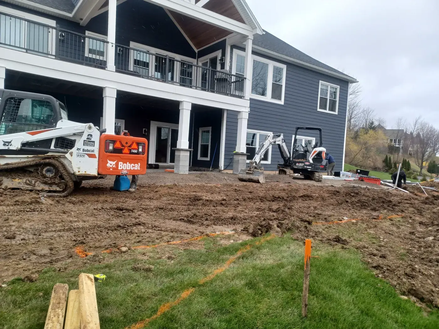 Backyard with the pool layout marked by orange stakes and a Bobcat and mini excavator on site before digging begins